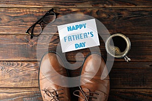 Brown leather shoes, inscription happy fathers day, cup of coffee and glasses on wooden background, space for text