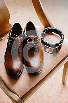 Brown men`s shoes made of genuine leather and a belt lie on a chair, close-up. Morning of the groom. Selective focus.