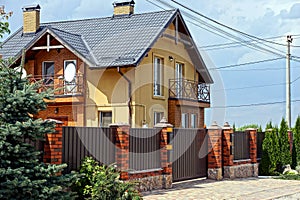 Brown metal fence with a gate in the street with decorative green trees in front of a large private house