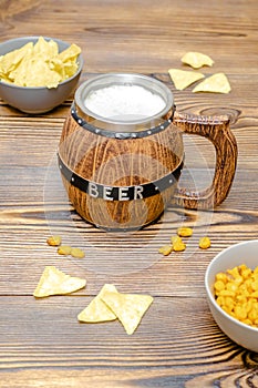 Brown mug of beer with foam and crispy snacks, pretzels, crackers, chips, nuts in bowl on wooden table, background close up.
