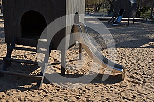 Brown painted wooden house on a playground with a sandpit. stainless steel slides shine in the sandy beach. wooden benches separat