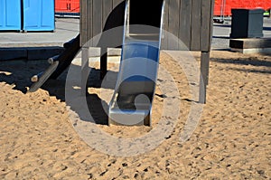 Brown painted wooden house on a playground with a sandpit. stainless steel slides shine in the sandy beach. wooden benches separat