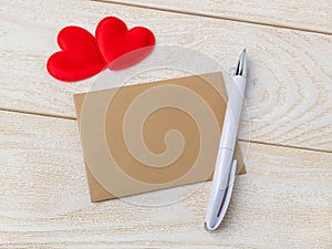 Brown paper empty greeting card, white ballpoint pen and two red silk valentine hearts on a white wooden textured background.