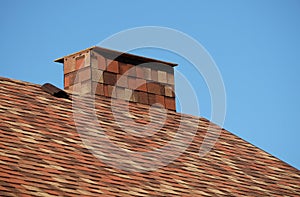 Brown roof of a house covered with soft shingles with short big smokestack close up under cloudless blue sky