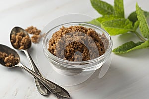 Brown sugar isolated in glass bowl, over styled marble background. Mint leaves. Top view, copy space