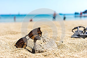 Brown sunglasses leaned towards a stone on a beach, flip flops in background