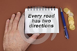 On a brown surface are coins, a pen and a notepad with the inscription - Every road has two directions