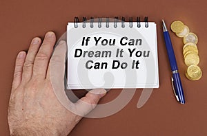 On a brown surface are coins, a pen and a notepad with the inscription - if You Can Dream It You Can Do It