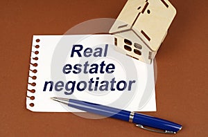 On a brown surface is a house, a pen and a notepad with the inscription - Real estate negotiator