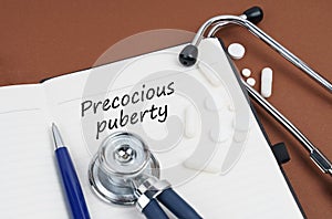 On a brown surface lie pills, a pen, a stethoscope and a notebook with the inscription - Precocious puberty