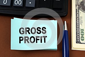 On a brown surface lies a calculator, dollars, pens and stickers with the inscription - gross profit