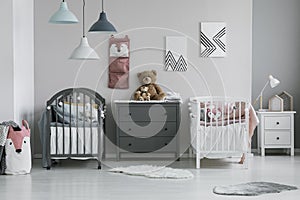 Brown teddy bear on grey wooden commode between two cribs in fashionable bedroom for twins