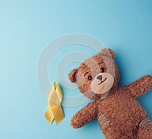 Brown teddy bear holds in his paw a yellow ribbon folded in a loop on a blue background. concept of the fight against childhood
