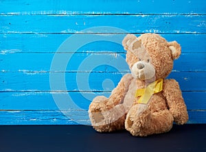 Brown teddy bear sits and a yellow silk ribbon on a blue wooden background, concept of the fight against childhood cancer