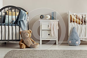 Brown teddy bear on the wooden floor of scandinavian baby bedroom interior with white nightstand and cribs