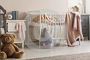 Brown teddy bear on the wooden floor of scandinavian baby bedroom interior with white nightstand and cribs