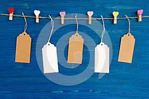 Blank paper price tags or labels set and wooden pins decorated on colored hearts hanging on a rope on the blue wooden background.