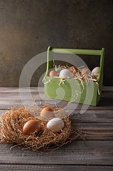 Brown and white chicken eggs in a straw nest on wooden background. Behind a wooden box of eggs in hay. Space under your