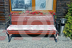 Brown wooden bench stands on the sidewalk near the wall of the house with a window and flowerpots with flowers