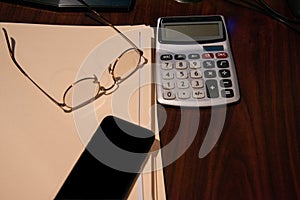 Brown wooden desk with several closed file folders a calculator and glasses turned upside down and a cell phone.