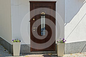 Brown wooden door on a gray wall and two flowerpots with flowers on the sidewalk on the street