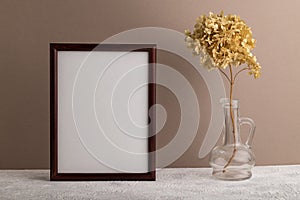 Brown wooden frame mockup with dried hydrangea in glass on beige paper background. Blank, vertical orientation, copy space