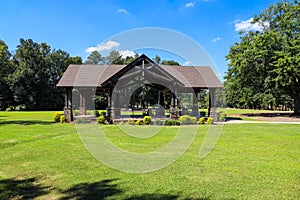 A brown wooden pergola near a like on vast green grass surrounded by lush green trees with blue sky and clouds