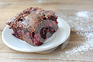 Brownie with cherries on a white plate. Close-up of chocolate cake on wooden table background