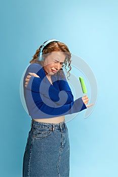 Brrr. Cold. Portrait of young girl in blue top, jeans skirt and headphones holding ice cream against blue studio photo