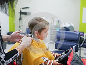 Brunet 2-3 years old boy sitting in big toy car at the hairdresser. Kid cut with hairdresser's machine