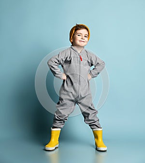 Brunet child in orange hat, gray overall, yellow rubber boots. He is smiling, hands on hips, posing on blue studio background