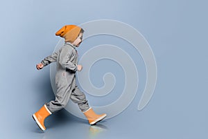 Brunet child in sunglasses, orange hat, gray overall and yellow rubber boots. He is running, posing on blue background