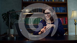Brunette 30s woman in glasses talking to laptop sitting at wooden table at home with bookcase. Working from home woman