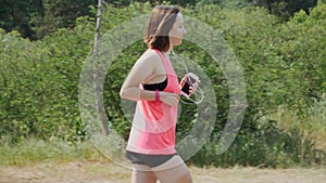 Brunette attractive girl in headphones and pink shirt running in park. Sportive woman training in park. Girl with funny sun tan ru