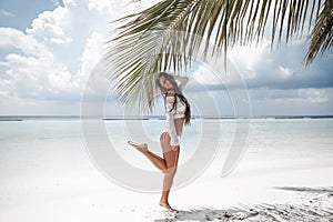 Brunette Bikini Model jumping by palm on sand on tropical beach vacation. Happy traveller woman. Sexy slim girl in white swimwear