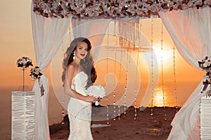 Brunette bride portrait. Wedding ceremony arch with flower arrangement and white curtain on cliff above sea at sunset, outdoor ph