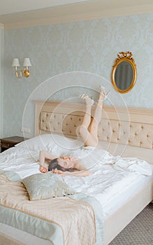 Brunette bride in wedding attire adorned with feathers and white high heels shoes close eyes, rest upside-down on bed.