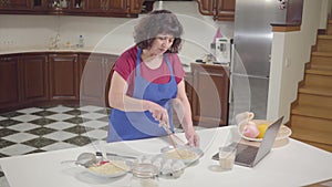 Brunette Caucasian mature woman cooking at the kitchen at home. Senior retiree checking recipe online and mixing dough