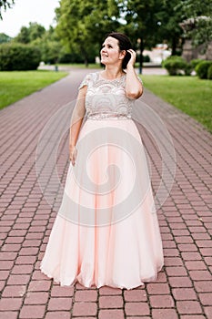 Brunette caucasian model in elegant floor-length pink dress dreamy looking away in park