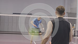 Brunette Caucasian sportsmen playing badminton in gym. Middle shot of confident athletic men training indoors. Live