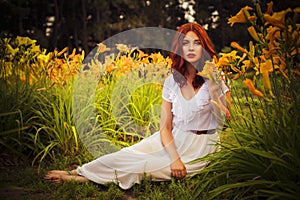 Brunette caucasian woman in white dress at the park in red and yellow flowers on a summer sunset holding flowers sitting on the gr