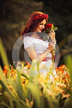 Brunette caucasian woman in white dress at the park in red and yellow flowers on a summer sunset holding roses