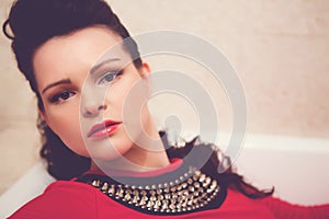 Brunette with curly hair in a red dress lies in an empty white bathtub