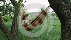 Brunette cute woman in hat and earphones listening to music, singing the song and using mobile phone in hammock. Charming female i