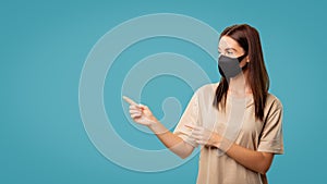 Brunette cute woman in mask pointing her fingers to copy space isolated over blue background