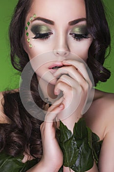 Brunette dryad woman with creative make up and beads on her face, curly hair and costume made of leaves on green backgro