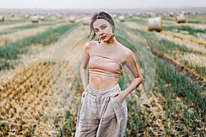 Brunette in fashionable clothes with bare shoulders standing in a wheat field on green grass. Woman holds her hands in pants