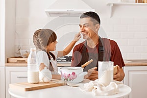 Brunette father wearing burgundy t shirt baking together with his female child in kitchen while sitting at table, daughter smeared