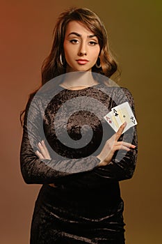 Brunette female in black velvet dress and jewelry showing two aces, posing against colorful studio background. Gambling