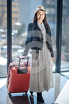 Brunette female in leather jacker and straw hat walking with red suitcase, smiling